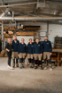 Sechs Personen in Arbeitskleidung stehen in einer Werkstatt mit Holzregalen und Maschinen.