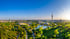 Luftaufnahme vom Olympiapark München mit Olympiaturm, Stadiondächern und See bei sonnigem Wetter.
