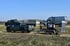 Grauer Pickup mit Anhänger und Minibagger, dahinter Transporter und LKW unter blauem Himmel.