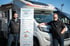 Zwei Mitarbeiter von Wohnmobile Harrislee posieren vor einem Roller Team Wohnmobil, flankiert von einem Informationsschild zu Serviceleistungen wie Vermietung und Verkauf.