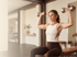 A woman in athletic wear sits on a weight bench performing an overhead shoulder press with two silver dumbbells.