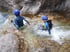 Zwei Personen in Neoprenanzügen und blauen Helmen sitzen im fließenden Wasser eines felsigen Flusses.