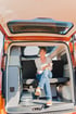 Lächelnde Frau in einem orangefarbenen Camper, die auf der Rückbank sitzt, umgeben von praktischen Stauräumen und einem klaren Blick auf die offene Heckklappe.