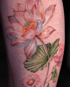 Ein farbiges Tattoo einer Lotusblüte mit rosa Blütenblättern, gelben Details und grünen Blättern auf heller Haut.