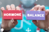 Hände halten rote und lila Blöcke mit den Worten "HORMONE" und "BALANCE" vor einem Molekülhintergrund.