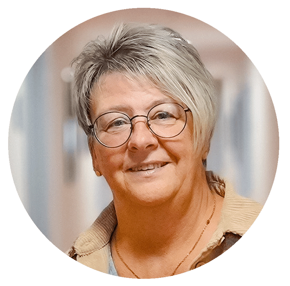 Eine lächelnde Frau mittleren Alters mit kurzen grauen Haaren und runder Brille.