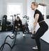 Zwei Frauen trainieren im Fitnessstudio; eine macht Kettlebell-Schwünge, die andere schwingt Trainingsseile.