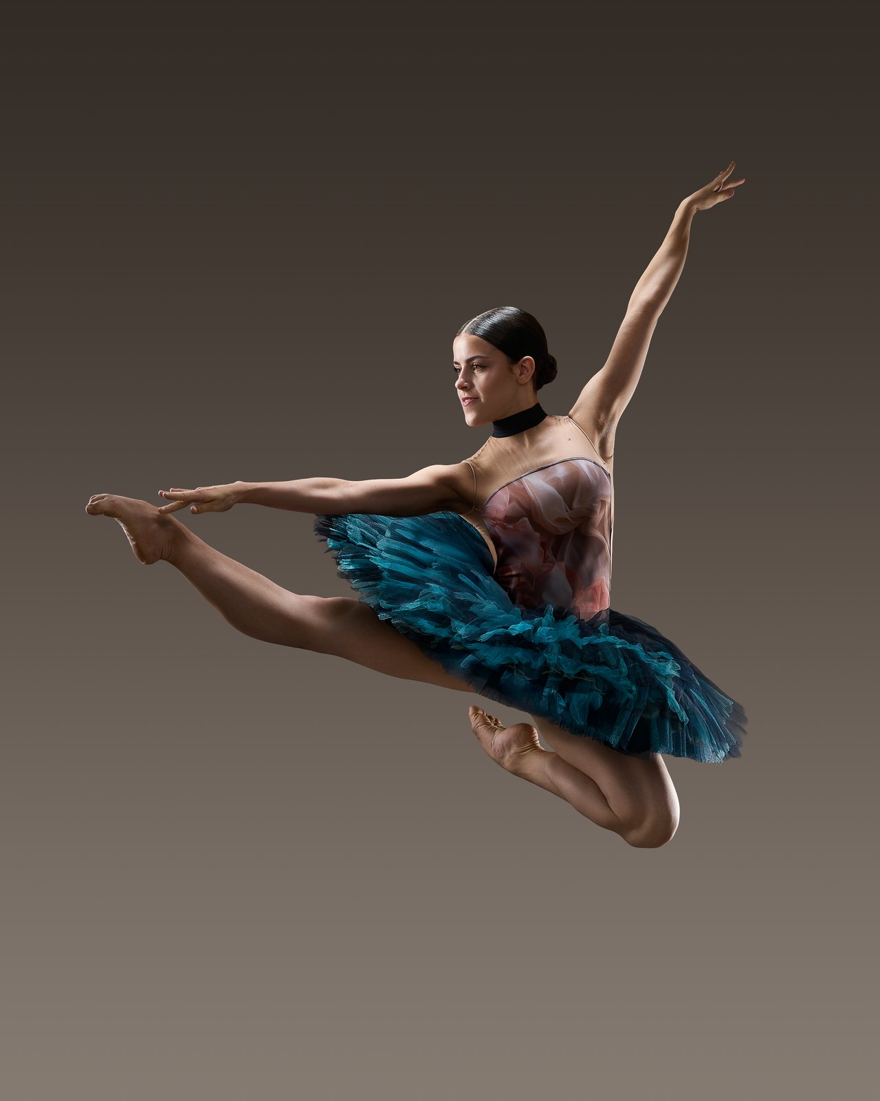 A dancer leaps in a blue-turquoise tutu against a warm grey background.
