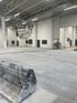 Eine leere Lagerhalle mit grauem Betonboden und weißen Wänden, im Vordergrund eine Baggerlade.