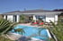 Moderner weißer Bungalow mit Swimmingpool, Terrasse, roten Liegestühlen und Palmen unter blauem Himmel.