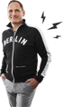 Ein lächelnder Mann in einer schwarzen Jacke mit "BERLIN"-Aufschrift und grauer Hose.