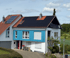 Haus mit roten Ziegeldächern, blauer Holzverkleidung und vielen schwarzen Solarmodulen auf den Dächern.