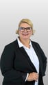 Sabine Eitner, professionelles Business-Porträt einer lächelnden Frau mit blonden Haaren, schwarzem Blazer und weißer Bluse vor grauem Hintergrund.