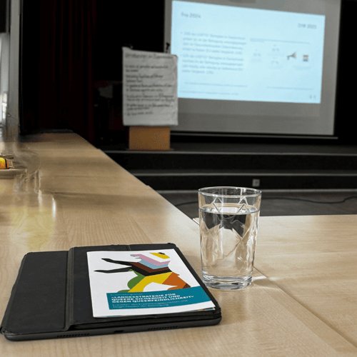 Tablet mit geöffnetem Buch, Wasserglas auf Holztisch, im Hintergrund eine Präsentation auf Leinwand.