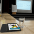 Ein Glas Wasser und ein Tablet mit bunter Broschüre liegen auf einem Holztisch vor einer Präsentationsbühne.