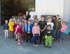 Gruppe von Kindern mit zwei Erwachsenen lächelt vor einer offenen Scheune mit Maschinen und Geräten.