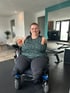 Eine lächelnde Frau mit kurzen Haaren und Brille sitzt in einem Elektrorollstuhl in einem modernen Raum.