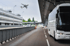 Ein weißer Reisebus parkt am Flughafen; ein Flugzeug fliegt im Hintergrund.