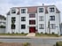 Modernes, weißes Mehrfamilienhaus mit roter Eingangstür, dunkelgrauen Fenstern und einer kleinen Rasenfläche davor.