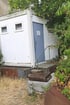Weißer Container mit blauer Tür und der Aufschrift "WC Damen", umgeben von trockenem Gras und Brombeersträuchern.