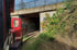 Roter Zug fährt auf Gleisen unter einer Betonbrücke hindurch, die Wände sind mit Graffiti bedeckt.