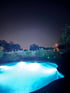 Beleuchteter Pool bei Nacht mit blauem Wasser, umgeben von einem Holzzaun, Gräsern und Bäumen.