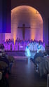 Ein Chor in blauen und weißen Gewändern singt vor einem Kruzifix, während eine Solistin vorne steht und Publikum im Vordergrund sitzt.