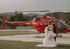 Brautpaar in Hochzeitskleidung steht vor einem roten Hubschrauber auf einem Flugplatz vor einer Hügellandschaft.