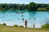 Badesee mit türkisfarbenem Wasser, vielen schwimmenden Menschen, Steg und bewaldetem Ufer.