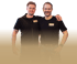 Zwei Männer in schwarzen T-Shirts, einer mit dem Arm um den anderen, lächeln in die Kamera.