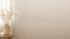 Hintergrundbild. Cremefarbene Wand, links ein leicht wehender lichtdurchlässiger heller Vorhang, davor eine cremefarbene Vase mit Trockenblumen.