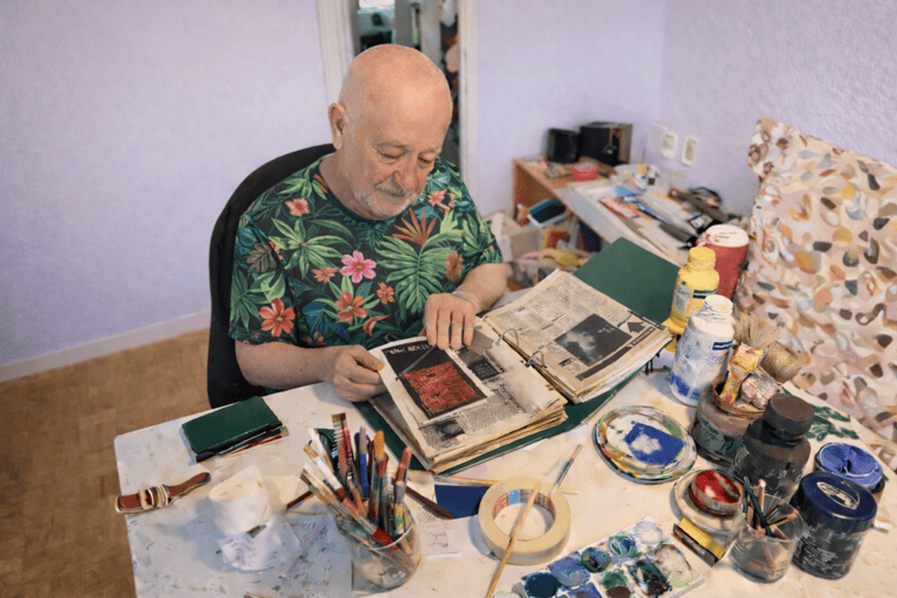 A man in a floral shirt sits at a cluttered desk, leafing through a notebook with newspaper articles.