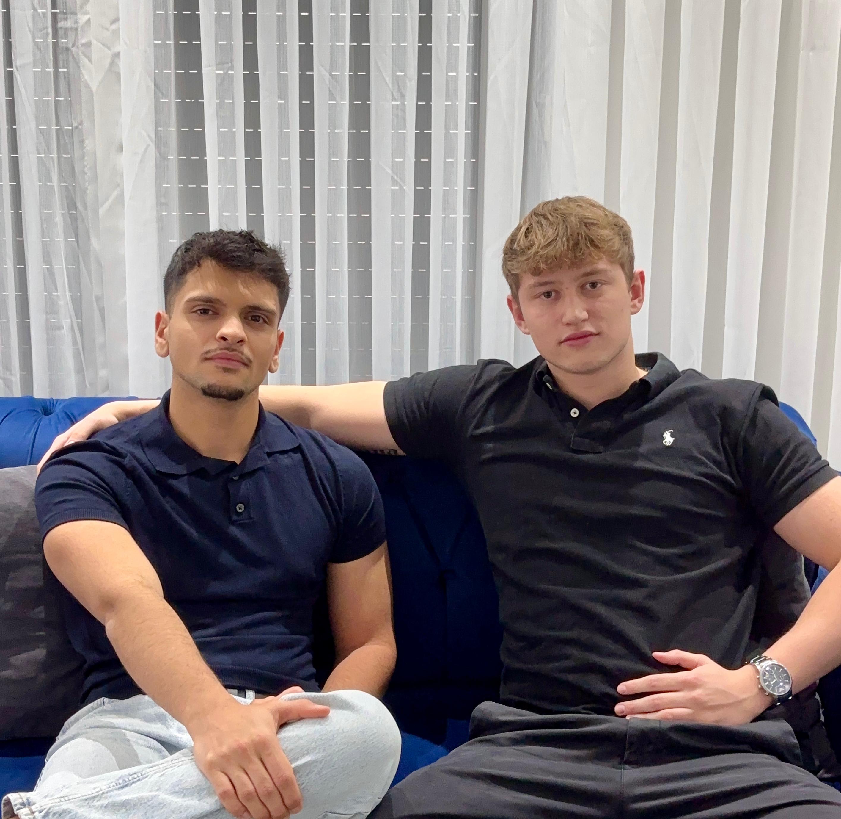 Zwei Männer sitzen auf einem blauen Sofa, einer mit dunklem Polo und Jeans, der andere mit schwarzem Polo und Hose.