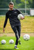 Mann in schwarzem Trainingsanzug hält einen Fußball und kickt einen anderen auf einem grünen Feld mit zwei weiteren Fußbällen.