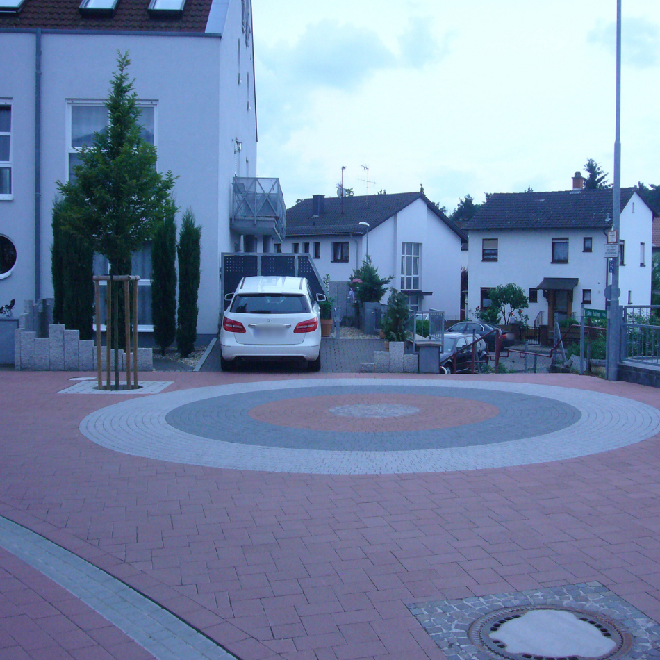 Ein weißes Auto parkt vor einem modernen Haus, davor ein kreisförmiges Pflastermuster in Rot, Grau und Weiß.