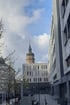 Ein Kirchturm mit Kuppel und spitzem Dach ragt zwischen modernen Gebäuden unter bewölktem Himmel hervor.