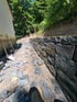 Neu gebaute Trockenmauer aus dunklen Natursteinen an einem Hang neben einem Haus mit Schaufel und Schutt im Vordergrund.
