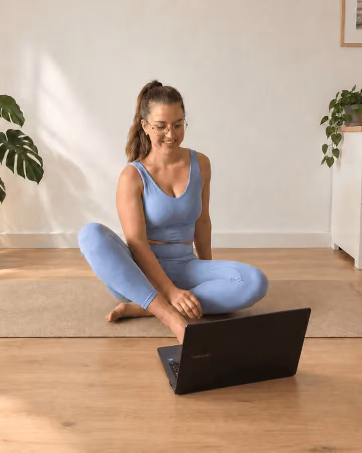 Online-Fitnesstrainerin Anna Maltan sitzt lächelnd auf der Yogamatte im Wohnzimmer vor ihrem Laptop und gibt ein Live-Online-Training
