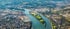 Luftaufnahme des Rheins, der an einer grünen Insel vorbeifließt und von einer Stadt mit Brücken und Häfen umgeben ist.