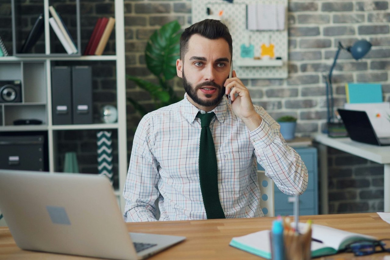 Mann in Hemd und Krawatte telefoniert mit dem Smartphone am Schreibtisch vor einem aufgeklappten Laptop im Büro
