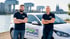 Zwei lächelnde Männer vor einem weißen Auto mit "Pflege am Rhein"-Logo und Kölner Skyline.