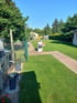 Gepflegter Garten mit grünem Rasen, gepflastertem Weg, Blumenkübeln und einer Schlauchtrommel unter blauem Himmel.