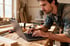 Carpenter in a workshop typing on a laptop with wood and tools on the workbench.