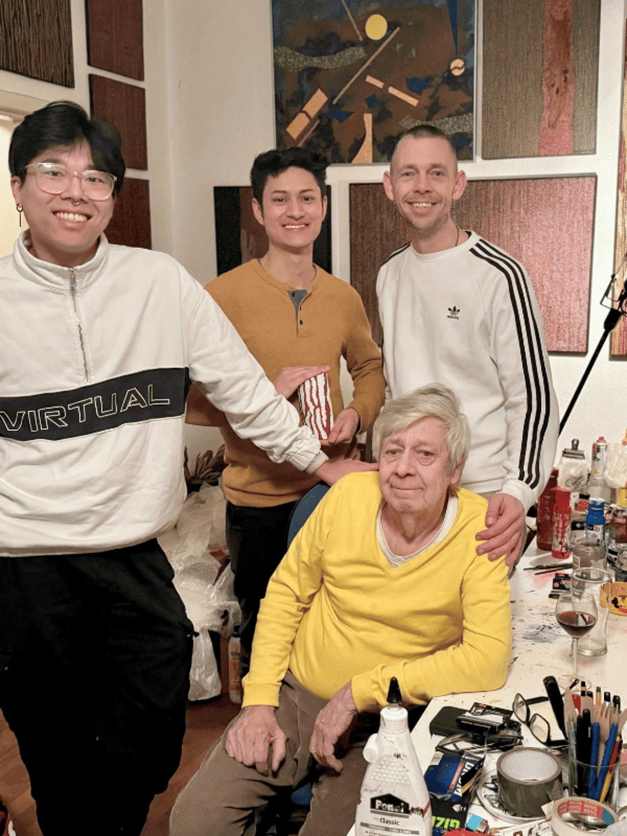 Four smiling men pose in front of abstract artworks. The oldest man sits at the front, the others stand.