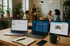 Drei Laptops und ein Notizbuch auf einem Holztisch mit verschwommenen Personen, die im Hintergrund diskutieren.