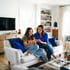 A woman and teenage girl sit on a white sofa in a bright living room, looking at a smartphone together.