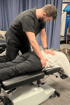 A male chiropractor in black scrubs performs a manual adjustment on a woman lying face down on a chiropractic drop table.