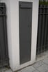 Graue Metallabdeckung mit Briefschlitz und zwei kleinen Klappen an einer weißen Mauer.