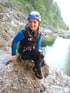Lächelnde junge Frau in Neoprenanzug und Helm hockt auf Felsen am Flussufer.
