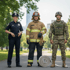 Polizistin, Feuerwehrmann mit Schlauch und Soldat in Uniform stehen auf einem Parkweg.